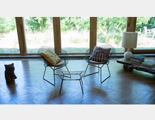 Wohnraum mit großen Holzfenstern, die Blick auf einen grünen Garten bieten. Im Vordergrund stehen zwei Stühle aus Drahtgeflecht mit Kissen, ein passender kleiner Tisch, sowie ein rustikaler Holztisch mit Stapeln von Büchern und einer Tischlampe.