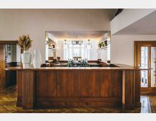 Barbereich mit einer Theke aus dunkelbraunem Holz, dekoriert mit weißen Vasen und getrockneten Pampasgräsern. Im Hintergrund sind Hängeregale mit Gläsern, eine Spiegelwand sowie große Fenster und moderne Hängelampen zu sehen.