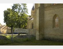Teilansicht einer Kirche aus hellem Backstein mit gotischen Fenstern, umgeben von Bauzäunen und einem Schild, das auf Baustellenbesichtigungen hinweist. Im Hintergrund sind Bäume und Wohnhäuser sichtbar.