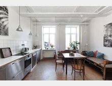 Helle Wohnküche mit modernen Edelstahlgeräten, weißen Fliesen, drei großen Fenstern und Pendelleuchten. Neben der Küchenzeile befindet sich eine Sitzecke mit Holztisch, Stühlen und einer rustikalen Holzbank mit farbigen Kissen.