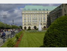 Ein Stadtplatz mit grüner Rasenfläche und Blumenbeeten im Vordergrund, einem zentralen Springbrunnen und dem historischen Hotel Adlon mit hellem, klassizistischem Fassadendesign im Hintergrund. Links eine Fußgängerzone mit Straßenlaternen und dem Berliner Fernsehturm in der Ferne.