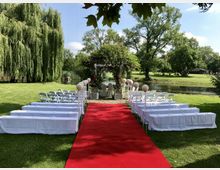 Eine Hochzeitskulisse im Freien mit einem roten Teppich, umgeben von weißen Stuhlreihen und geschmückten Ständern. Im Hintergrund ein blühender Pavillon, Bäume, eine Weide und ein Teich auf einer grünen Wiese.