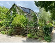 Einfamilienhaus mit grau verputzter Fassade, teilweise bewachsen mit Efeu, umgeben von einem Garten mit hoher Vegetation und einer niedrigen Holzgitterzaunbegrenzung; Gehweg und gepflasterte Straße im Vordergrund.