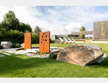 Ein gepflegter Garten mit einer modernen Zierinstallation aus rostfarbenem Metall mit floralen Ausschnitten, umgeben von Kies und Rasenflächen. Im Hintergrund stehen Liegestühle auf einer Holzterrasse und ein Gebäude mit klaren Linien aus Holz und Glas.