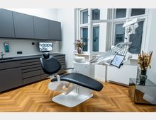 Moderner Zahnarzt-Behandlungsraum mit einem schwarzen Behandlungsstuhl, einem weißen Instrumentenarm mit Bildschirm, grauen Schränken und hellem Fischgrätenparkettboden. Im Hintergrund sind ein Waschbecken, ein Fenster mit Milchglas sowie ein Blumenarrangement auf einem kleinen Tisch sichtbar.