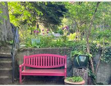 Pinkfarbene Gartenbank vor einer niedrigen Steinmauer, dahinter ein üppig begrünter Garten mit lila Tulpen, Bäumen und Sträuchern; daneben ein großer grüner Blumentopf auf einem Holzstamm. Eine steinerne Treppe mit schwarzem Geländer führt links nach oben.