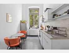 Schmale, helle Küche mit großem Fenster am Ende, das Blick auf grüne Bäume bietet. Auf der linken Seite befinden sich ein runder Esstisch mit zwei orangefarbenen Stühlen und ein Kühlschrank, auf der rechten Seite eine Küchenzeile mit Spüle, Herd und offenen Regalen für Geschirr.