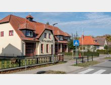 Ein Gebäude der Freiwilligen Feuerwehr (FFW) mit cremefarbener Fassade, roten Fensterrahmen und einem Ziegeldach, daneben weitere Wohnhäuser im ähnlichen Baustil. Im Vordergrund befinden sich ein Fußgängerüberweg, eine Zaunanlage und Straßenschilder.