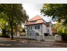 Zweistöckiges Einfamilienhaus mit hellgrauer Fassade, roten Dachziegeln und blauen Fensterläden, umgeben von einem weißen Gartenzaun. Das Gebäude steht an einer gepflasterten Straße mit großen, schattenspendenden Bäumen.