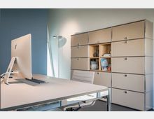 Modernes Büro mit minimalistischem Schreibtisch, der einen großen Monitor trägt, und einem weißen Bürostuhl. Im Hintergrund steht ein beige-farbener, modularer Aktenschrank mit offenen und geschlossenen Fächern, daneben eine schlanke Stehleuchte.