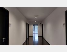 Flur eines modernen Bürogebäudes mit weißen Wänden, dunklem Bodenbelag und schwarzen Türrahmen. Am Ende des Flurs befindet sich ein Fenster mit vertikalen Jalousien, das Tageslicht hereinlässt.