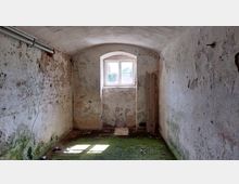 Kleiner, leerer Raum mit bröckelnden Putzwänden und einem gewölbten Deckenbogen. Ein einfaches Fenster im hinteren Bereich lässt Tageslicht herein, der Boden ist mit Moos und Schmutz bedeckt.