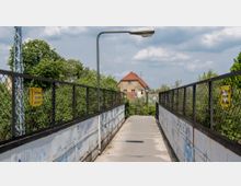 Fußgängerbrücke mit Metallgittern an den Seiten, begleitet von gelben Warnschildern mit der Aufschrift „Hochspannung Lebensgefahr“. Im Hintergrund ist ein Wohnhaus mit rotem Dach, umgeben von Bäumen, zu sehen.