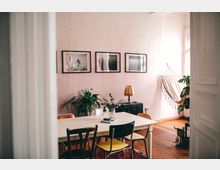 Ein gemütlicher Wohnraum mit Holzboden und einer weißen Wand, an der drei gerahmte Schwarz-Weiß-Bilder hängen. Im Mittelpunkt steht ein weißer Tisch mit unterschiedlichen Stühlen, daneben Pflanzen, eine Tischlampe auf einer Truhe und eine cremefarbene Hängematte am Fenster.