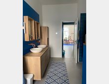 Badezimmer mit blauer Akzentwand, Doppelwaschbecken mit weißen Schalen auf einer Holzkonsole sowie Kupferarmaturen. Im Hintergrund führt eine offene Tür in einen hellen Raum mit Pflanzen auf einem Tisch.