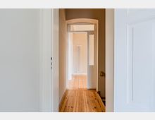 Schmaler Innenflur in einer Wohnung mit hellem Holzdielenboden, hohen weißen Türrahmen und einem Rundbogen am Ende des Gangs. Durch eine offene Tür ist ein angrenzender Raum mit hohem, schmalem Heizkörper an der Wand zu sehen.