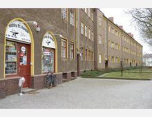 Außenansicht eines Wohnhauskomplexes mit brauner Fassade und gelben Fensterrahmen, links im Bild ein Geschäft mit der Aufschrift „Hobby-RC-Hangar“ und Schaufenstern. Vor dem Geschäft steht ein Fahrrad, und im Vordergrund liegt ein gepflasterter Gehweg, angrenzend an eine Grünfläche mit einzelnen Bäumen.