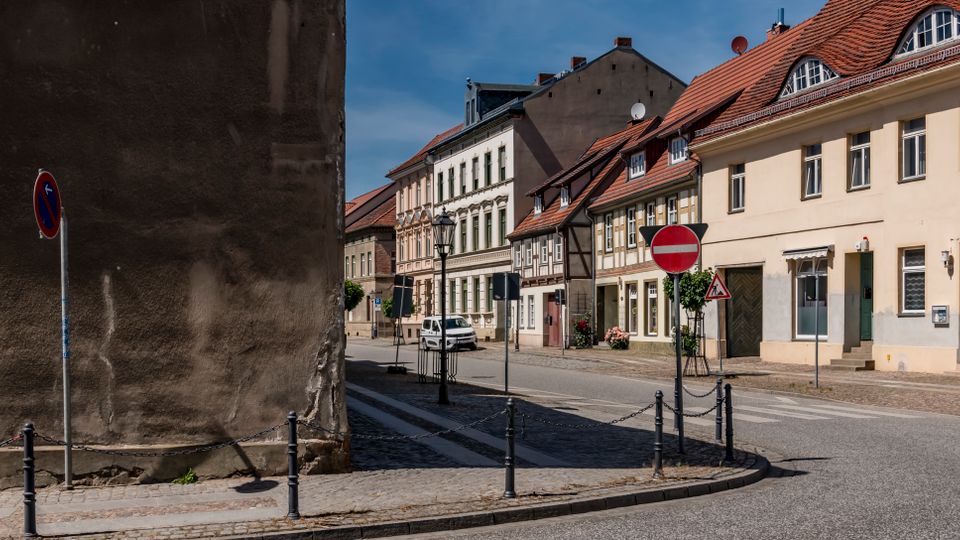 Straßenecke in einer historischen Altstadt mit Kopfsteinpflaster und einer Reihe von Wohnhäusern mit Putzfassaden, teils Fachwerk und roten Ziegeldächern. Im Vordergrund stehen Verkehrsschilder und Poller mit Ketten, links dominiert eine hohe, fensterlose Brandwand.