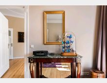 Ein elegantes Interieur mit einem kleinen Beistelltisch aus dunklem Holz und Marmorplatte, auf dem ein dekoratives Glas mit blauen Schmetterlingen steht. An der Wand hängt ein goldgerahmter Spiegel, der teilweise den Raum reflektiert, und im Hintergrund sind Holzdielen sowie ein Türdurchgang erkennbar.