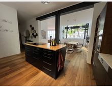 Moderne Wohnküche mit offenem Essbereich, ausgestattet mit einem zentralen Küchenblock aus Holz und schwarzen Schubladen. Im Hintergrund befindet sich ein Esstisch mit Stühlen vor einer großen Fensterfront, die viel Tageslicht hereinlässt.