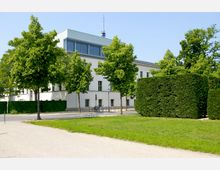 Ein modernes weißes Gebäude mit großen Fenstern im oberen Stockwerk, umgeben von Bäumen und gepflegten Hecken. Im Vordergrund befindet sich eine Rasenfläche, ein gepflasterter Weg sowie ein Fahrradständer.