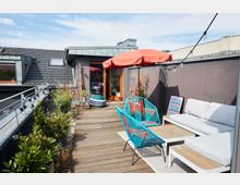 Dachterrasse mit Holzboden, modernen Gartenmöbeln in Weiß und Türkis, einem roten Sonnenschirm, Pflanzen entlang einer Brüstung und Lichterkette. Im Hintergrund ein Gebäude mit dunkler Fassade und Fenstern im Dach.