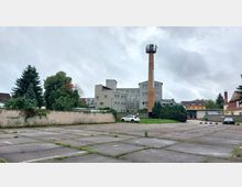 Leerer Parkplatz mit vereinzeltem Grasbewuchs, umgeben von einer niedrigen Ziegelmauer. Im Hintergrund stehen ein mehrstöckiges graues Gebäude mit großen Fenstern, ein hoher Backsteinschornstein mit Plattform, Bäume sowie weitere kleinere Gebäude.