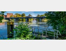 Szene eines Flusses mit einer Metallbrücke im Hintergrund, über die Fahrzeuge fahren, und einem kleinen Steg im Vordergrund, umgeben von dichtem Grün. Im Hintergrund ist ein Backsteingebäude sowie Bäume entlang des Ufers zu sehen.