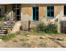 Eine alte Hausfassade mit bröckelndem Putz und blauen Fensterrahmen, daneben ein hölzernes Tor. Im Vordergrund befindet sich ein mit Gras und Erde bewachsener, trockener Hof, vor dem Gebäude stehen Pflanzen und ein Kaminofen.