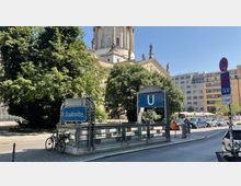 U-Bahn-Eingang "Stadtmitte" mit verzierten Metallgeländern, umgeben von Bäumen und einem Teil eines historischen Gebäudes im Hintergrund. Daneben eine Straße mit parkenden Autos und modernen Gebäuden im städtischen Umfeld.