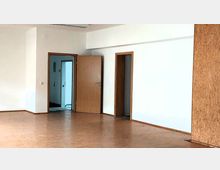 Leerer Innenraum mit hellem Laminatboden, weiß gestrichenen Wänden und drei Türen mit Holzrahmen, von denen eine teilweise geöffnet ist und in einen angrenzenden Raum führt.