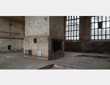 Industriehalle mit hohen Wänden aus Ziegelsteinen, großen Fenstern mit Metallrahmen und einem zentralen gemauerten Schornstein. Der Raum wirkt verlassen, der Fußboden ist beschädigt, und durch die Fenster ist spärliche Vegetation im Außenbereich sichtbar.