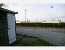 Eine Außenaufnahme mit einem kleinen Holzschuppen links im Vordergrund, einer gepflasterten Fläche und einer Hecke in der Mitte. Im Hintergrund sind Flutlichtmasten und ein Gebäudekomplex zu sehen.