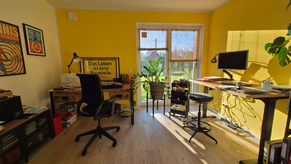 Helles Arbeitszimmer mit gelb gestrichener Wand, einem höhenverstellbaren Schreibtisch, Bildschirm und Bürostuhl. Große Fenster im Hintergrund mit Blick ins Freie, Zimmerpflanzen und Wanddekoration ergänzen die Einrichtung.