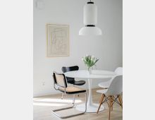 Minimalistisch eingerichtetes Esszimmer mit einem weißen runden Tisch, umgeben von verschiedenen Designerstühlen auf hellem Holzboden. An der Wand hängt ein abstraktes Linienbild, daneben eine moderne, weiße Pendelleuchte und eine Vase mit weißen Blumen auf dem Tisch.