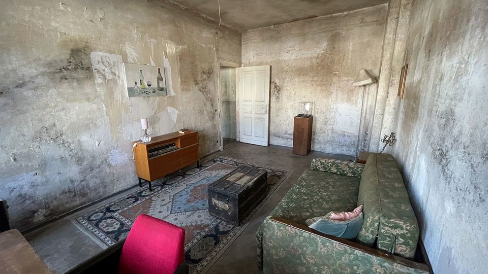 Innenraum eines heruntergekommenen Wohnzimmers mit stark fleckigen, abblätternden Wänden und einer offenen weißen Tür. Im Raum stehen ein grünes gemustertes Sofa, ein Orientteppich, eine Truhe als Couchtisch sowie ein niedriger Holzschrank und einzelne Lampen.