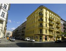 Ecke eines Wohnviertels mit einem großen, leuchtend gelben Altbau mit reich verzierten Fassadenelementen und Balkonen, im Erdgeschoss befindet sich ein Gasthaus. Kopfsteinpflasterstraßen, geparkte Autos und weitere mehrstöckige Gebäude säumen die Straße, unter blauem Himmel mit spärlichen Wolken.