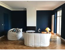 Wohnzimmer mit dunkelblauen Wänden, Parkettboden und modernen, weißen Sitzmöbeln. Im Raum befinden sich ein Beistelltisch, eine Stehlampe mit orangefarbenem Schirm und ein Fenster mit Glastür, das Tageslicht hereinscheinen lässt.