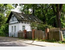 Weißes Fachwerkhaus mit schwarzem Dach und Dachflächenfenstern, umgeben von hohen Bäumen. Vor dem Haus befindet sich ein Zaun aus Metall und Holz mit gemauerten Backsteinsäulen entlang einer gepflasterten Straße.