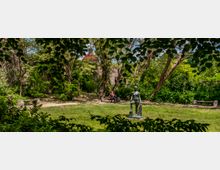 Grüne Parklandschaft mit Bäumen und einer Rasenfläche, im Vordergrund eine moderne Bronze-Skulptur. Im Hintergrund sind eine historische Steinmauer, eine Bank mit einer Person und ein rotes Gebäude mit einem Turm zu sehen.