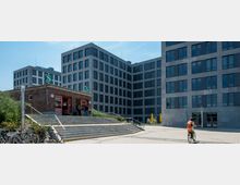 Backsteingebäude eines S-Bahnhofs mit zwei grünen "S"-Schildern, umgeben von modernen, grauen Bürogebäuden mit vielen Fenstern; davor befinden sich eine Treppe, Fahrräder und eine Person auf einem Fahrrad.