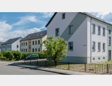 Wohngebiet mit mehrstöckigen Wohnhäusern in Pastellfarben, flankiert von gepflegten Hecken und einem Baum im Vordergrund. Im Hintergrund steht ein geparktes Auto, während ein Gehweg und eine Straße das Bild ergänzen.