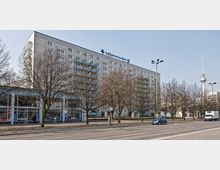 Wohngebäude mit mehreren Balkonen an einer breiten Straße, flankiert von unbelaubten Bäumen. Im Hintergrund ist der Berliner Fernsehturm sichtbar.