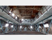 Industriehalle mit offenliegender Holzkonstruktion und Metallträgern. Die Wände sind aus Backstein, es gibt Fenster auf zwei Ebenen, und eine umlaufende Galerie erstreckt sich entlang der oberen Ebene.