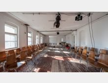 Ein simpler, heller Raum mit Holzboden und weiß gestrichenen Wänden, bestückt mit zwei Reihen aus Holzstühlen auf beiden Seiten. An der Decke sind Scheinwerfer und Kabel angebracht, während Sonnenlicht durch mehrere Fenster an der linken Wand einfällt.