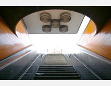 Treppe und Rolltreppenaufgang in einer U-Bahn-Station mit orange gefliesten Wänden und runden Lüftungselementen an der Decke. Am oberen Ende der Treppe scheint helles Tageslicht hinein.
