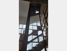 Verlassene Treppe in einem Gebäude mit großen, vergitterten Fenstern und sichtbaren Metall- und Holzelementen; die Umgebung wirkt unfertig oder im Verfall begriffen.