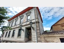 Zweistöckiges historisches Gebäude mit Putzfassade, teils freigelegtem Mauerwerk und dekorativen Fensterrahmen. Vor dem Eingang befinden sich zwei Laternen an schmiedeeisernen Halterungen, daneben ein Anbau mit rotem Ziegeldach.