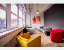 Heller Aufenthaltsraum mit großen Fenstern, grauer Wand und modernem Designmöbelmix, darunter graue Sofas, ein gelber und ein roter Sessel. An der Wand hängt ein abstraktes Gemälde in Rot- und Orangetönen, der Raum wird durch eine Hängelampe beleuchtet.