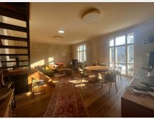 Wohnraum mit Holzfußboden und großen, bodentiefen Fenstern, die viel Tageslicht hereinscheinen lassen. Im Vordergrund ein runder Esstisch mit Stühlen, im Hintergrund eine gemütliche Sitzecke mit Sofas und dekorativen Kissen, dazu eine Holztreppe und ein Teppich.